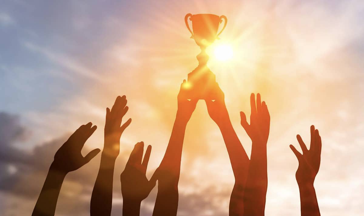 Multiple hands holding a trophy up (with the sun shining behind their hands)