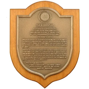 Shield-shaped bronze historical discovery plaque mounted on a solid wood backing featuring a scientific atom logo.